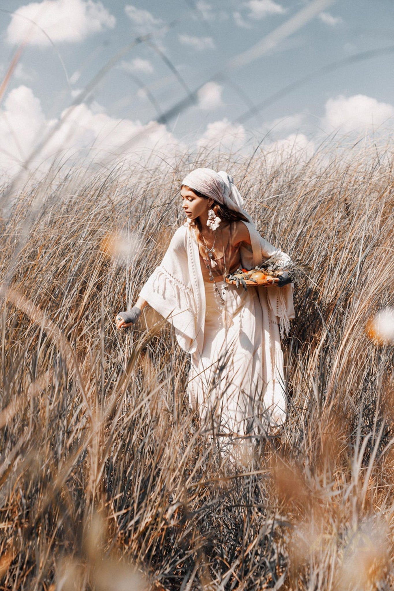 A woman dressed in a white dress and shawl gracefully walks through a field of tall grass, wearing the AYA Sacred Wear's Boho Kimono Cardigan complemented by a stylish headscarf. She gazes downward while holding a bouquet of flowers, as the sky, dotted with scattered clouds, enhances the serene atmosphere.