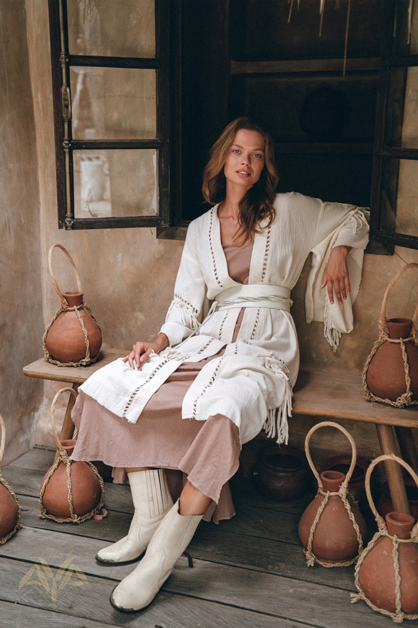 A woman in a Boho Kimono Cardigan by AYA Sacred Wear sits on a wooden bench, surrounded by decorative pottery. The background features an open window with rustic wooden shutters. She is wearing beige boots and a light dress underneath the cardigan.