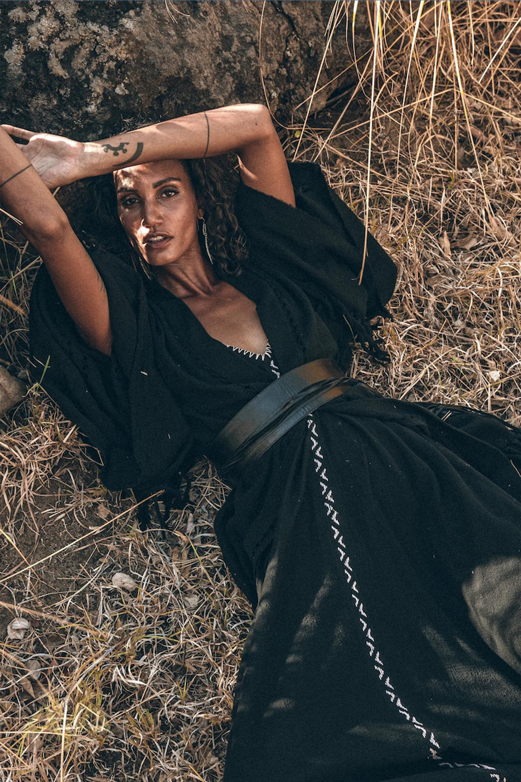 A woman reclines on dry grass, leaning against a rock with her arms raised. She wears a black dress featuring white detailing and a wide belt, paired beautifully with AYA Sacred Wear's Boho Kimono Cover Up. Sunlight casts shadows on her face, enhancing her serene expression.