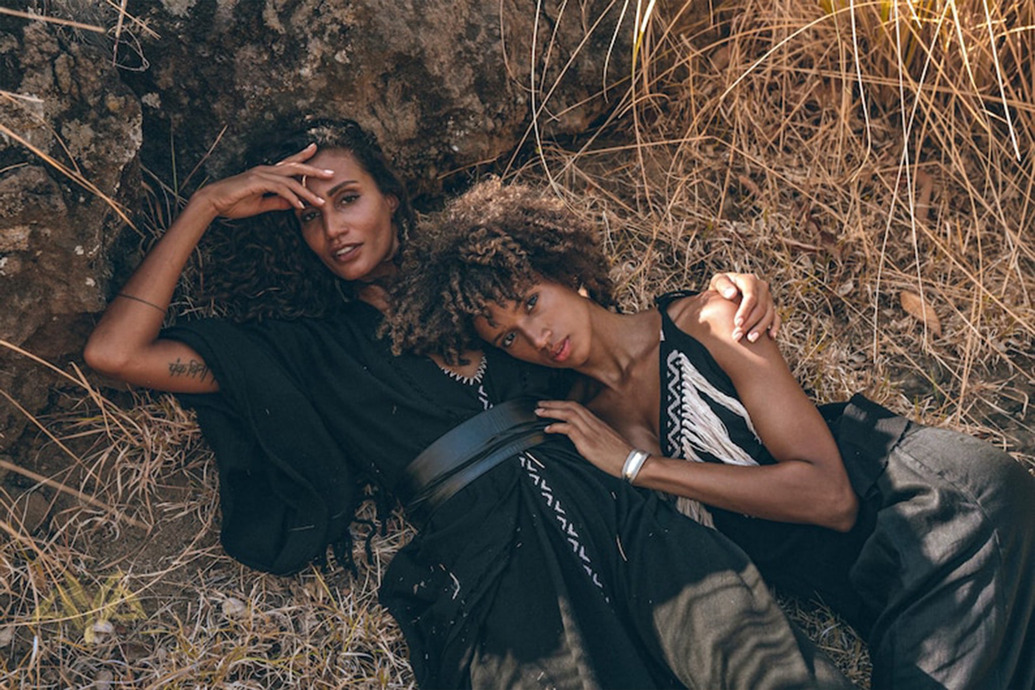 Two people lie on the ground against a rocky backdrop surrounded by dry grass. They are dressed in black, patterned clothing, including an AYA Sacred Wear Boho Kimono Cover Up with wide sleeves. Both appear relaxed and content, with one person's head resting on the other's shoulder.