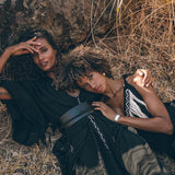 Two people lie on the ground against a rocky backdrop surrounded by dry grass. They are dressed in black, patterned clothing, including an AYA Sacred Wear Boho Kimono Cover Up with wide sleeves. Both appear relaxed and content, with one person's head resting on the other's shoulder.