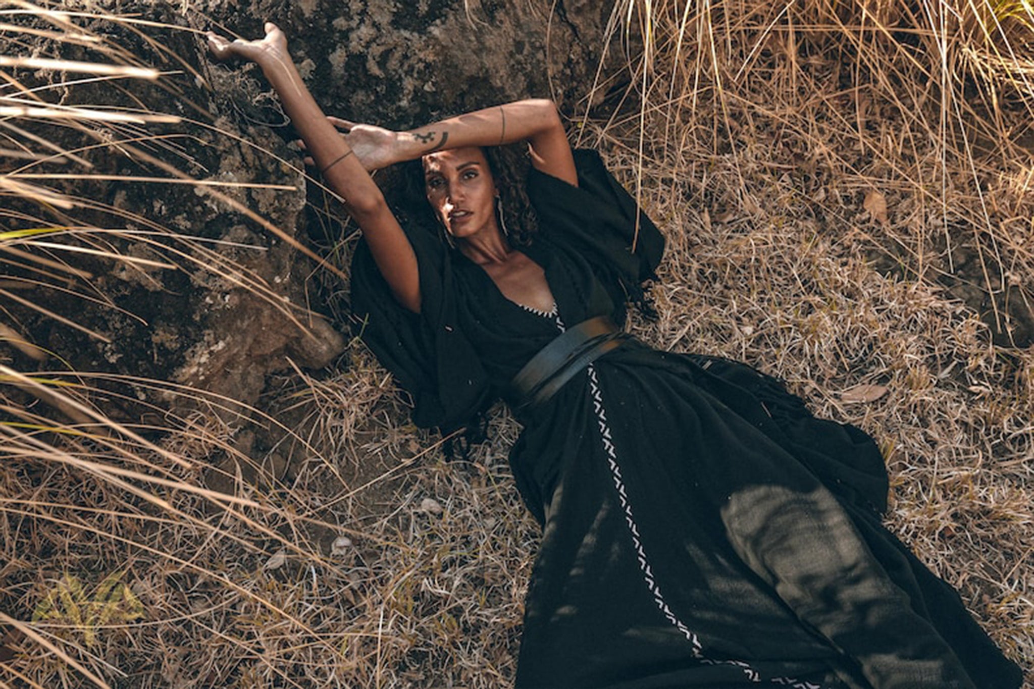 A person wearing a long black dress and the Boho Kimono Cover Up from AYA Sacred Wear, featuring wide sleeves and a cardigan mantle style, lies on dry grass, leaning against a rock. They have one arm extended over their head and the other behind it, surrounded by tall, dry grass and plants.