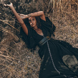 A person wearing a long black dress and the Boho Kimono Cover Up from AYA Sacred Wear, featuring wide sleeves and a cardigan mantle style, lies on dry grass, leaning against a rock. They have one arm extended over their head and the other behind it, surrounded by tall, dry grass and plants.