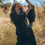 A person with curly hair elegantly poses among tall grass, draped in the Boho Kimono Cover Up—a wide sleeve cardigan mantle from AYA Sacred Wear. The background showcases a clear blue sky and a distant mountain.