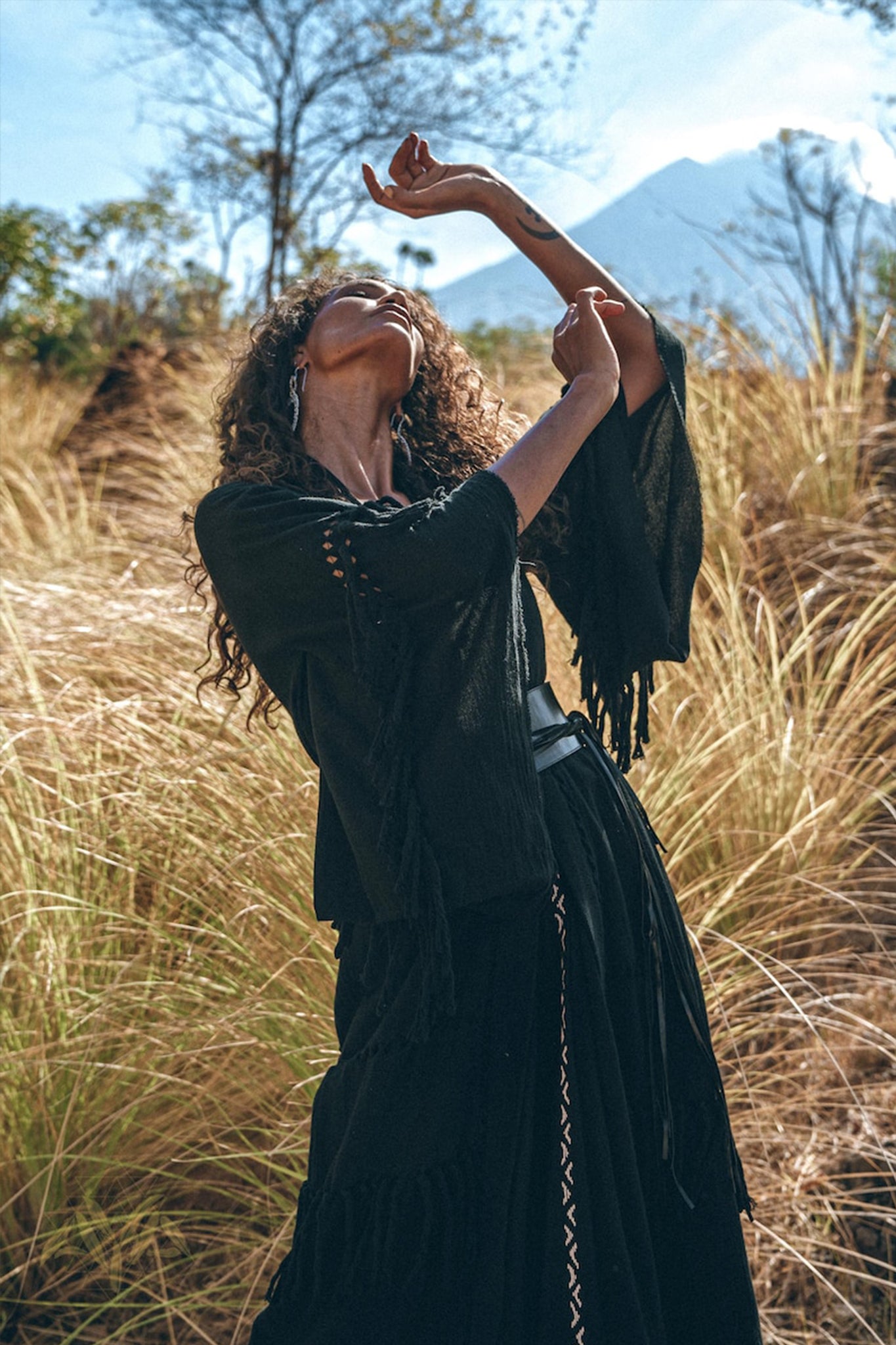 A person with curly hair elegantly poses among tall grass, draped in the Boho Kimono Cover Up—a wide sleeve cardigan mantle from AYA Sacred Wear. The background showcases a clear blue sky and a distant mountain.