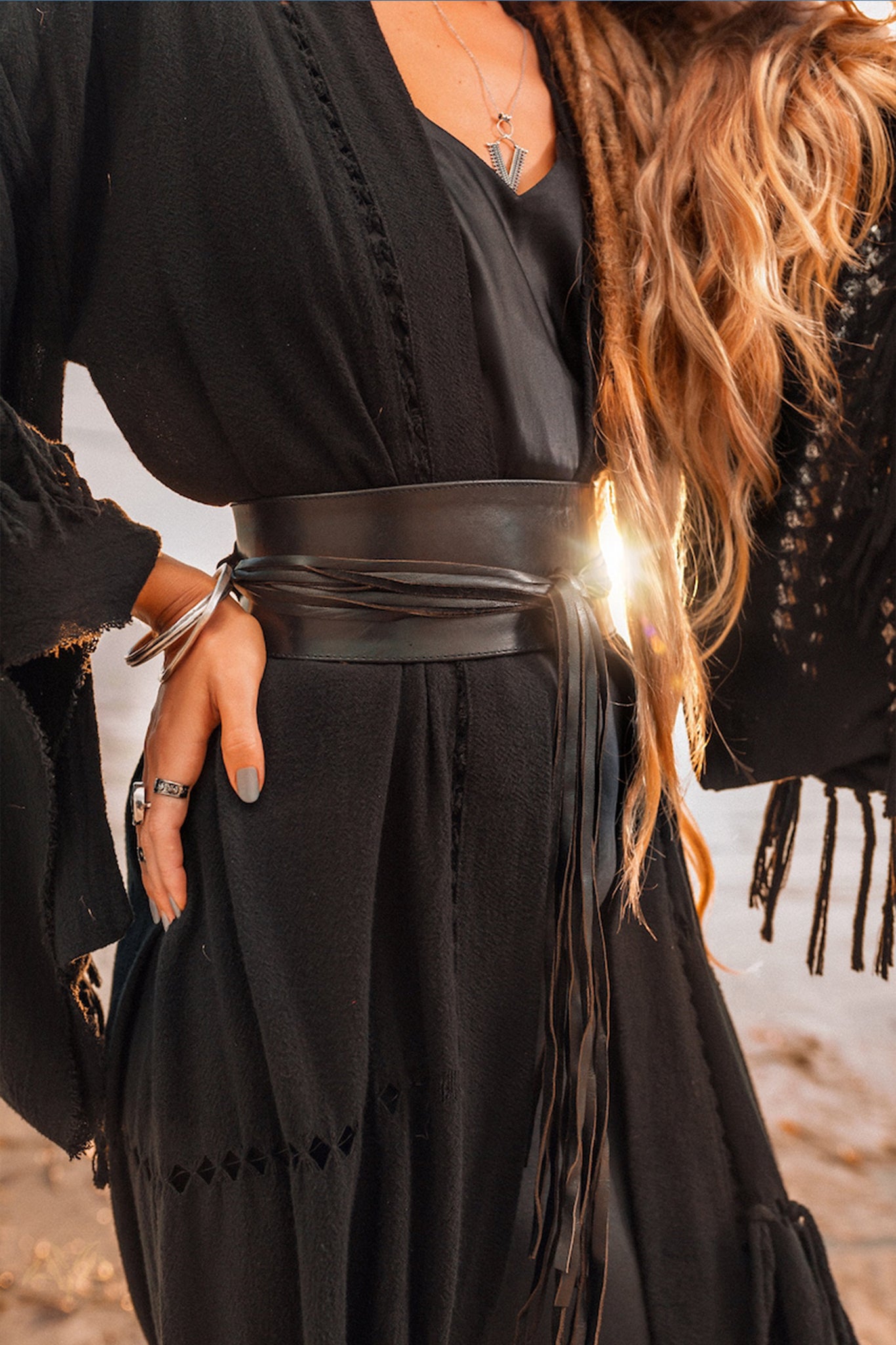 Standing outdoors with sunlight glimmering off their silver bangles and rings, a person in a black outfit with a wide leather belt and long hair embodies bohemian style. This essence is accentuated by the flowing Boho Kimono Cover Up from AYA Sacred Wear, featuring wide sleeves and serving as the perfect cape for women. Resting a hand on their hip completes their stylish look.