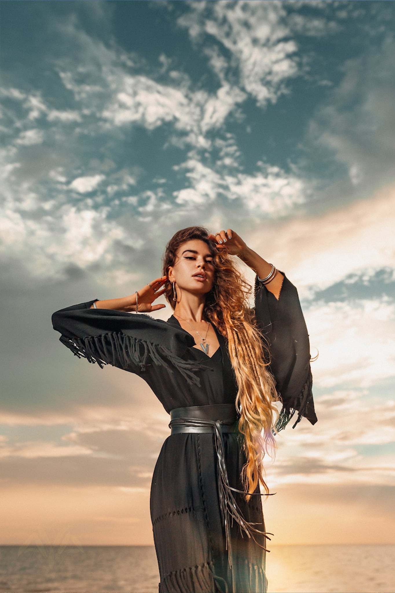 A person with long hair stands on a beach at sunset, draped in an AYA Sacred Wear Boho Kimono Cover Up with wide sleeves. Their pose is confident, with one hand touching their hair and the other behind their head. The partly cloudy sky adds a dramatic effect to the scene.