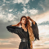 A person with long hair stands on a beach at sunset, draped in an AYA Sacred Wear Boho Kimono Cover Up with wide sleeves. Their pose is confident, with one hand touching their hair and the other behind their head. The partly cloudy sky adds a dramatic effect to the scene.
