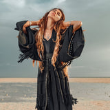 Under the dramatic, overcast skies, a woman in a flowing black dress stands on the beach with eyes closed and hands in her hair. She exudes calm and confidence, as though wrapped in an invisible Boho Kimono Cover Up by AYA Sacred Wear, effortlessly blending into the serene natural scenery.