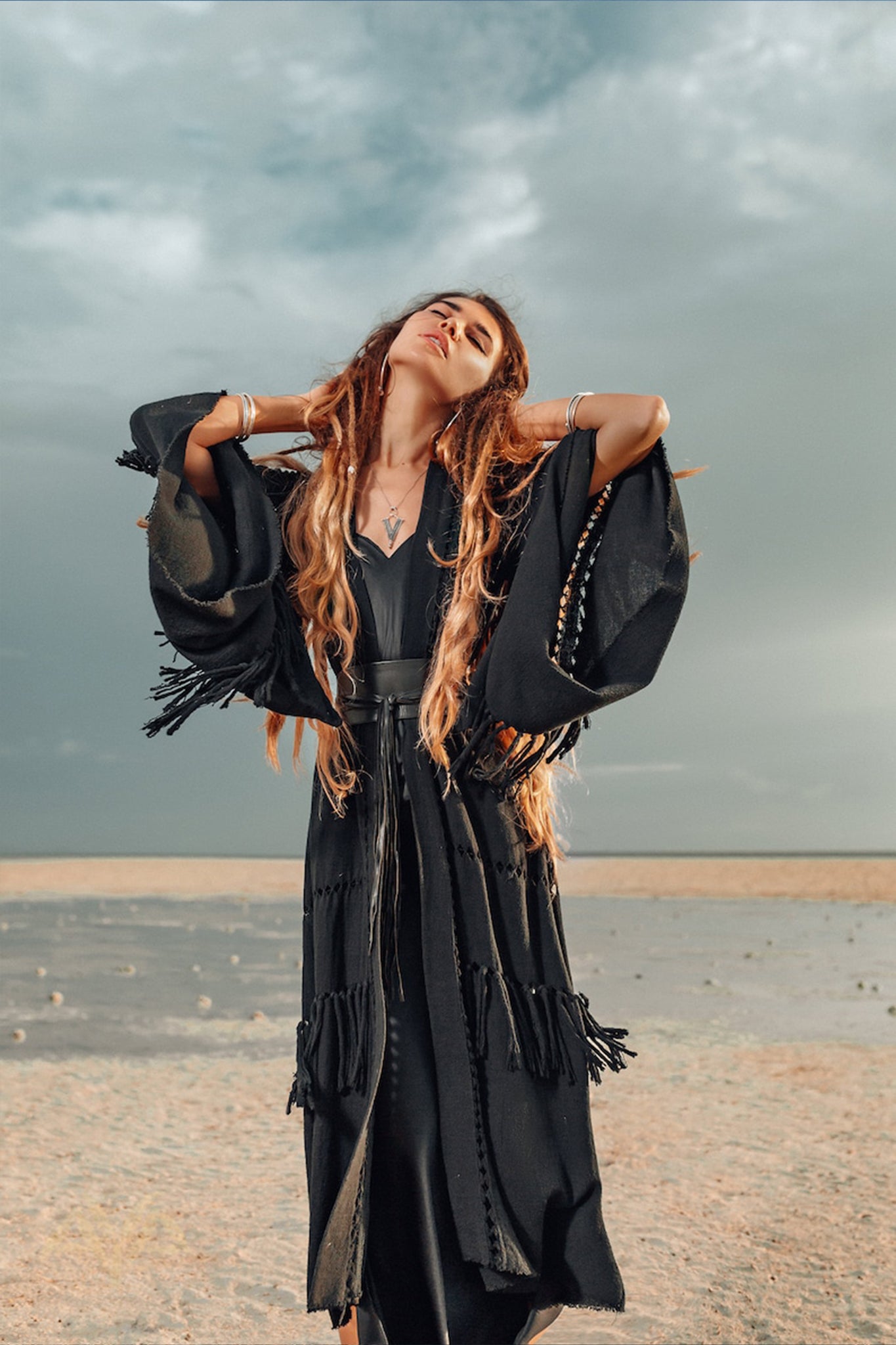 Under the dramatic, overcast skies, a woman in a flowing black dress stands on the beach with eyes closed and hands in her hair. She exudes calm and confidence, as though wrapped in an invisible Boho Kimono Cover Up by AYA Sacred Wear, effortlessly blending into the serene natural scenery.