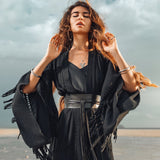 A woman with long hair stands on a beach, wearing the Boho Kimono Cover Up, a wide sleeve cardigan mantle by AYA Sacred Wear, along with several bracelets. Her eyes are closed as she looks upward. The background reveals a cloudy sky and sandy shore, perfectly complementing her fringe-embellished cape crafted from natural cotton.