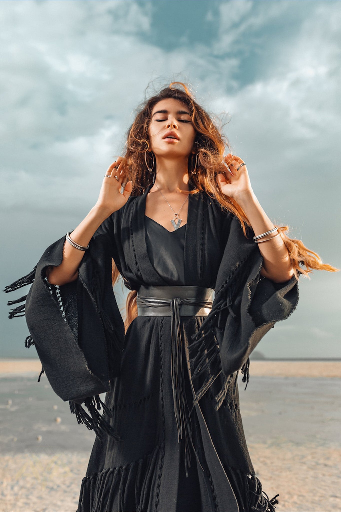 A woman with long hair stands on a beach, wearing the Boho Kimono Cover Up, a wide sleeve cardigan mantle by AYA Sacred Wear, along with several bracelets. Her eyes are closed as she looks upward. The background reveals a cloudy sky and sandy shore, perfectly complementing her fringe-embellished cape crafted from natural cotton.