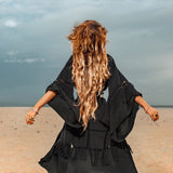 A person with long, wavy hair strides across a vast, sandy landscape beneath a cloudy sky, adorned in an AYA Sacred Wear Boho Kimono Cover Up. Its wide sleeves and flowing design evoke a sense of freedom and solitude.