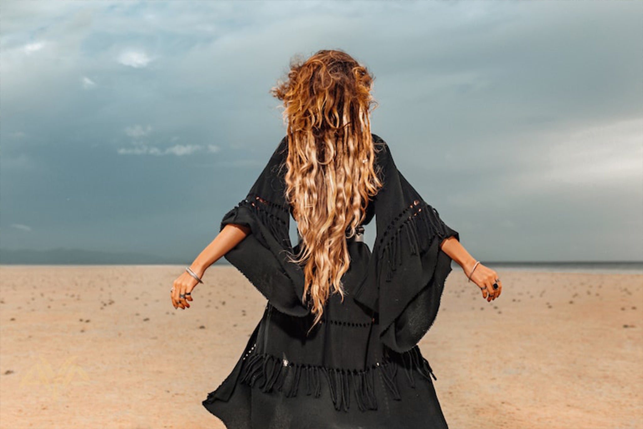 A person with long, wavy hair strides across a vast, sandy landscape beneath a cloudy sky, adorned in an AYA Sacred Wear Boho Kimono Cover Up. Its wide sleeves and flowing design evoke a sense of freedom and solitude.