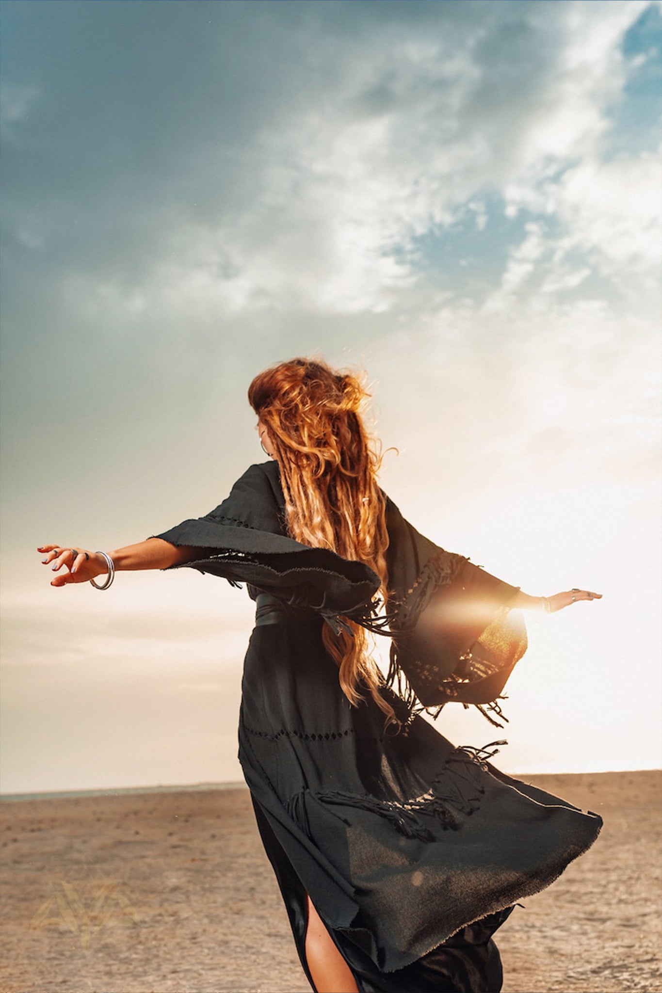 A person with long hair wearing a black flowing dress and an AYA Sacred Wear Boho Kimono Cover Up—featuring wide sleeves and a cardigan mantle—dances joyfully on a beach at sunset. The partly cloudy sky and the sun's warm glow create a serene atmosphere, perfectly complementing the bohemian vibe.