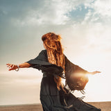 A person with long hair wearing a black flowing dress and an AYA Sacred Wear Boho Kimono Cover Up—featuring wide sleeves and a cardigan mantle—dances joyfully on a beach at sunset. The partly cloudy sky and the sun's warm glow create a serene atmosphere, perfectly complementing the bohemian vibe.