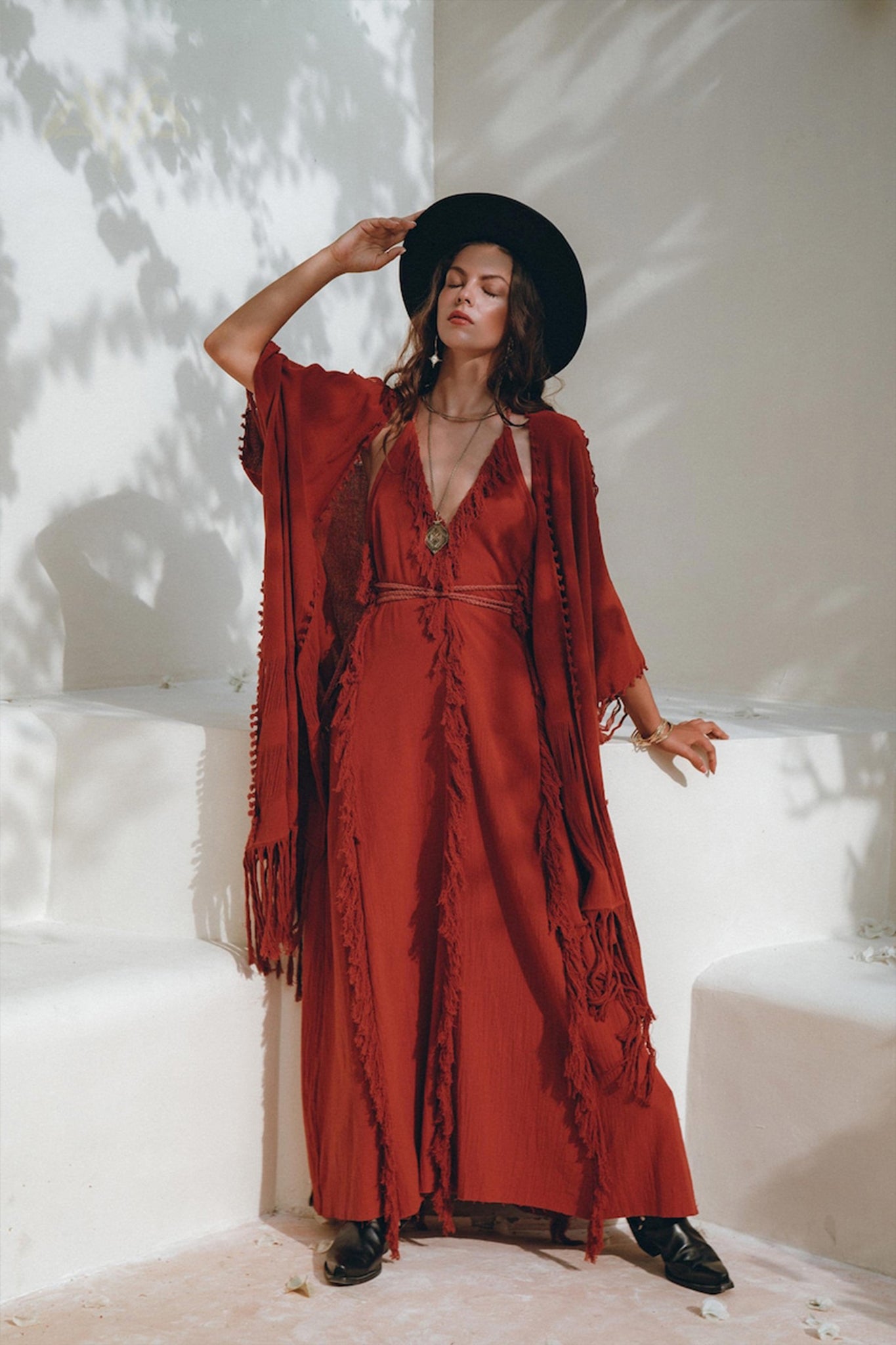 A person in a long, AYA Sacred Wear Boho Maxi Dress, also known as the Red Organic Goddess Dress or Cotton Bridesmaid Dress, complemented by a wide-brimmed hat stands against a light-colored wall. They are adorned with layered necklaces and bracelets, with sunlight casting soft shadows around them.