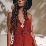 A woman with long wavy hair wears a black hat and a sleeveless red dress from AYA Sacred Wear. The dress, known as the "Boho Maxi Dress • Cotton Bridesmaid Dress • Red Organic Goddess Dress," features fringe details. She stands in front of a neutral background with her eyes closed, basking in dappled sunlight and shadow patterns.