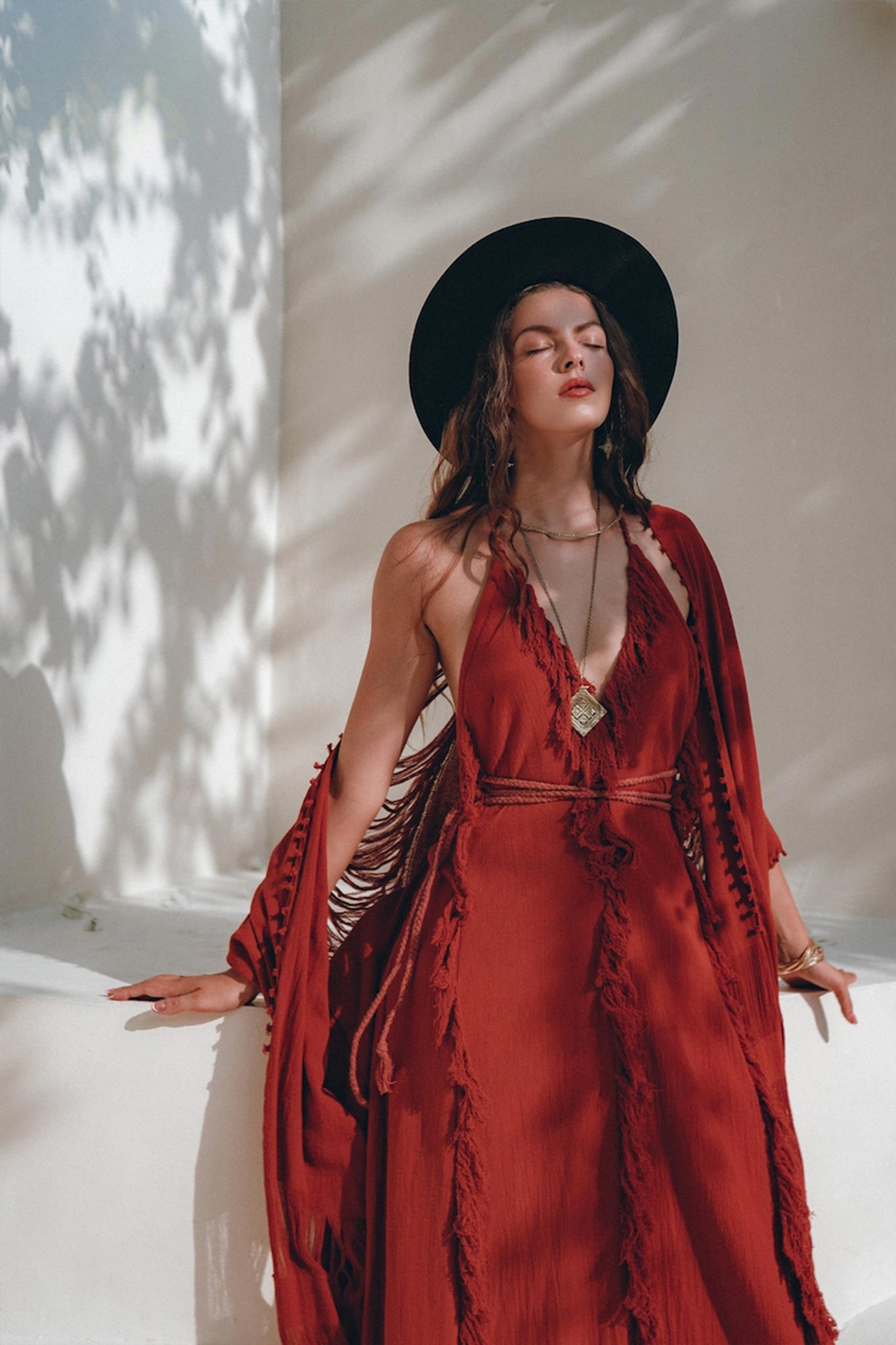 A woman wearing the Red Organic Goddess Dress by AYA Sacred Wear, complemented with a black hat, stands against a sunlit wall. With eyes closed, she savors the sunlight as leaf shadows create patterns across the wall. She looks relaxed, with one hand gently resting on the surface beside her.