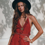 A woman with long, wavy hair is wearing a wide-brimmed black hat and the Boho Maxi Dress by AYA Sacred Wear. This sleeveless red organic goddess dress features fringe details and she stands outdoors against a light background dappled with shadow patterns, accessorized with a necklace and bracelets.