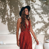 A woman in a flowing Red Organic Goddess Dress by AYA Sacred Wear and a black hat stands outdoors. She is surrounded by white walls and vases, with sunlight casting shadows of leaves on the wall behind her.