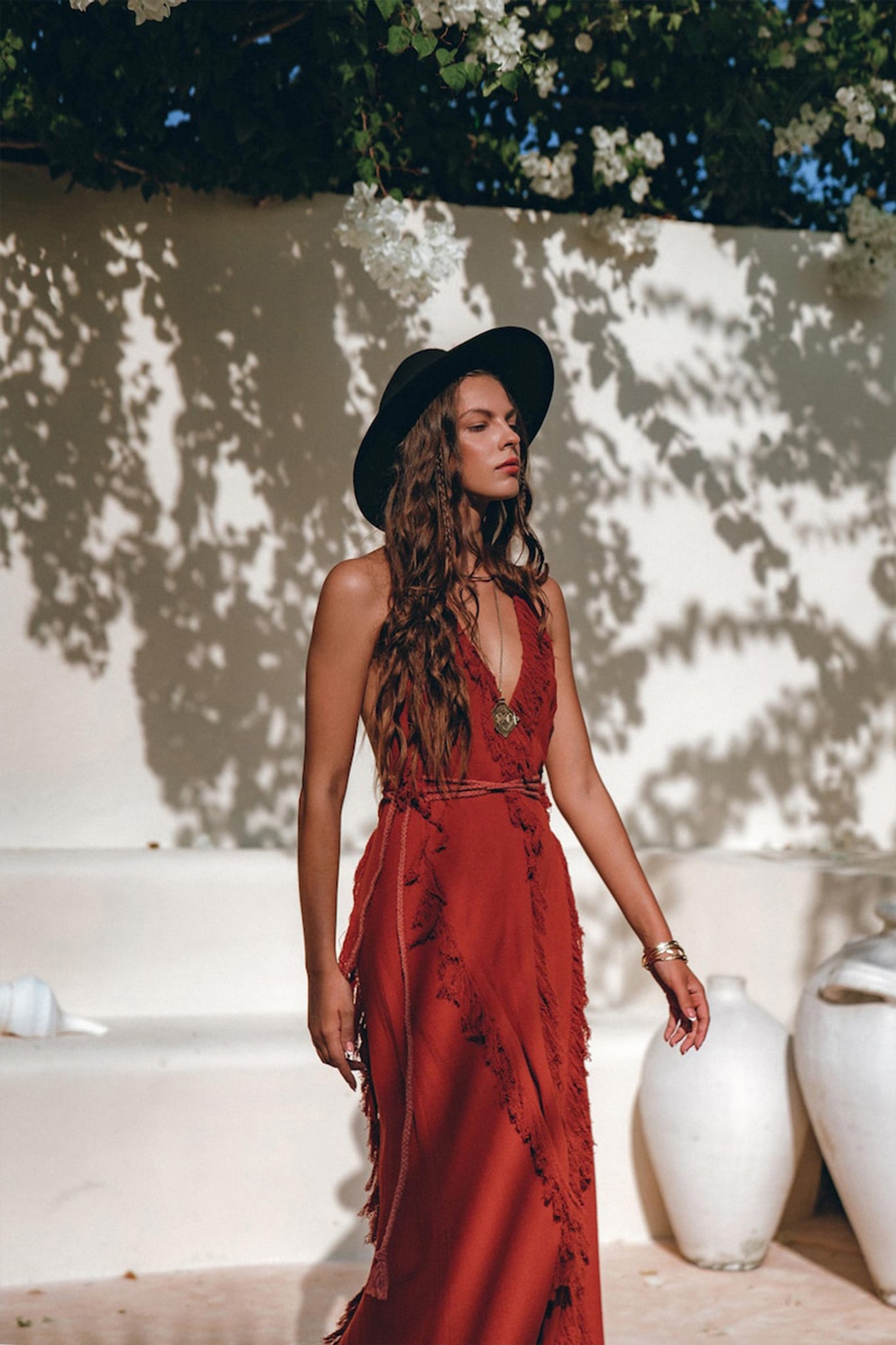 A woman in a flowing Red Organic Goddess Dress by AYA Sacred Wear and a black hat stands outdoors. She is surrounded by white walls and vases, with sunlight casting shadows of leaves on the wall behind her.