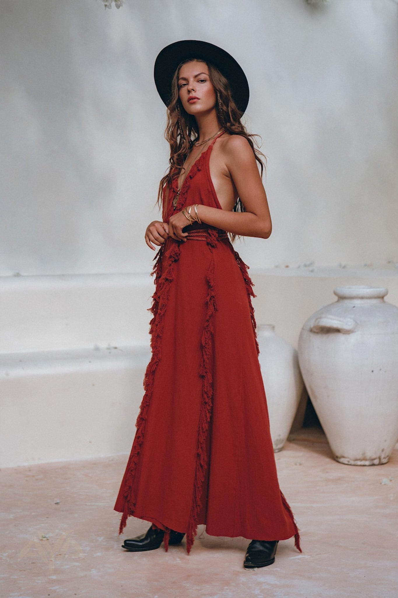 A woman exudes confidence in a Red Organic Goddess Dress, a long, sleeveless boho maxi piece by AYA Sacred Wear. She pairs it with a black hat and boots, her long wavy hair cascading around her. The scene is set against white walls complemented by large ceramic pots.