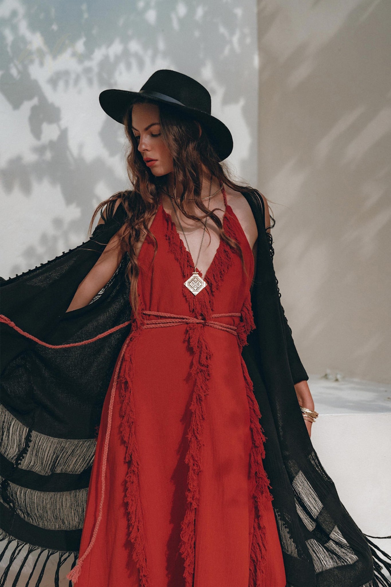 A woman poses outdoors in a chic red dress from AYA Sacred Wear, paired with a black hat. Her look is enhanced by a black shawl and a long necklace. Sunlight creates shadows on the wall behind her, providing an enchanting backdrop for her Boho Maxi Dress ensemble.