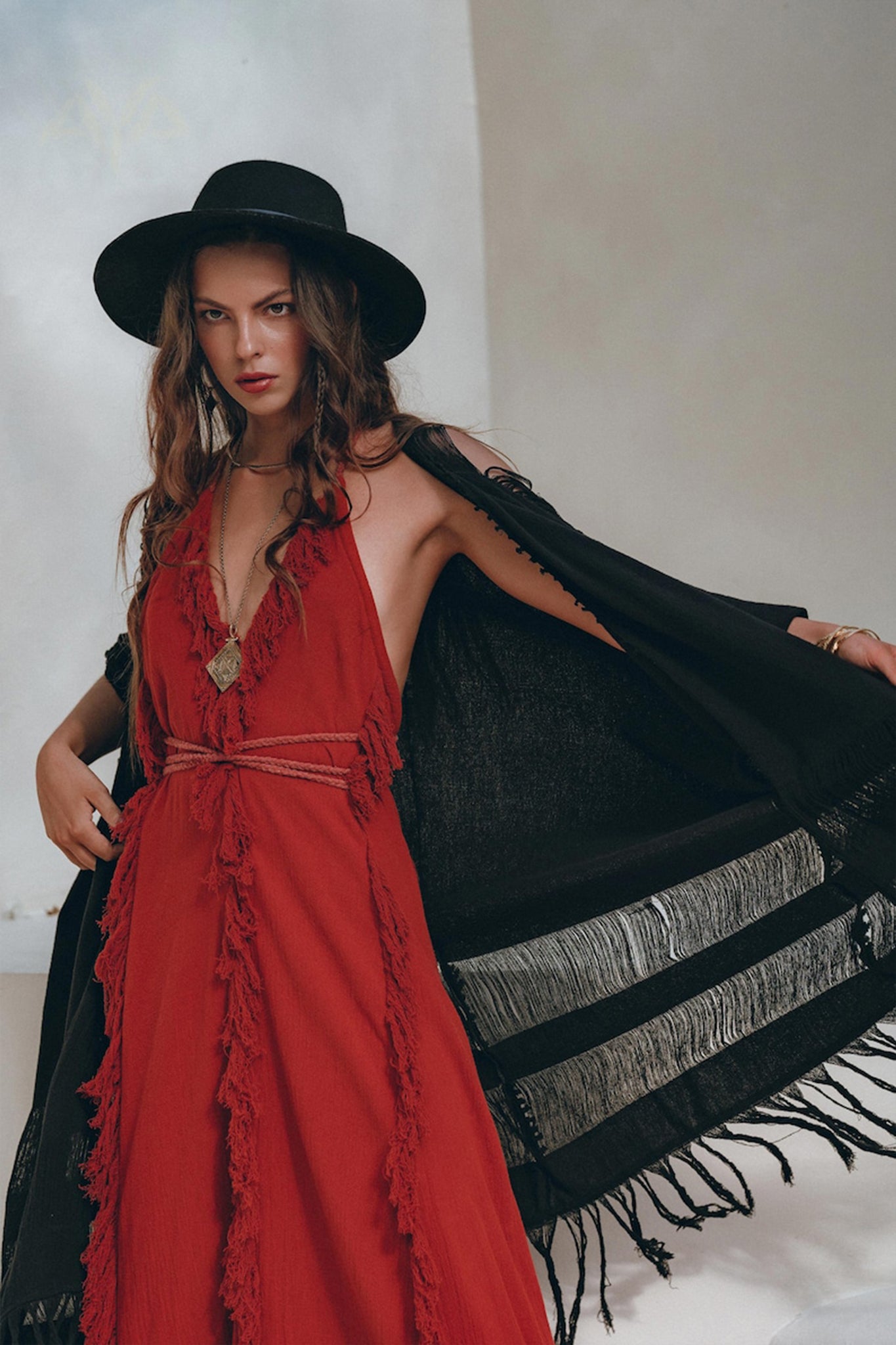 A woman exudes confidence as she poses in a flowing red organic goddess dress by AYA Sacred Wear. Her bohemian style is further enhanced by a black wide-brimmed hat and a black shawl draped over her shoulders, with fringes catching the light on one side. The neutral backdrop accentuates the organic elegance of her outfit.