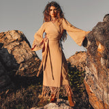 A woman with long, wavy hair stands on rocky terrain, exuding confidence in a tan, long-sleeved dress and boots. She embraces the spirit of ethical fair work production while posing against the rocks under a clear sky, wearing the Boho Maxi Skirt by AYA Sacred Wear — a sustainable cotton skirt that embodies bohemian elegance.