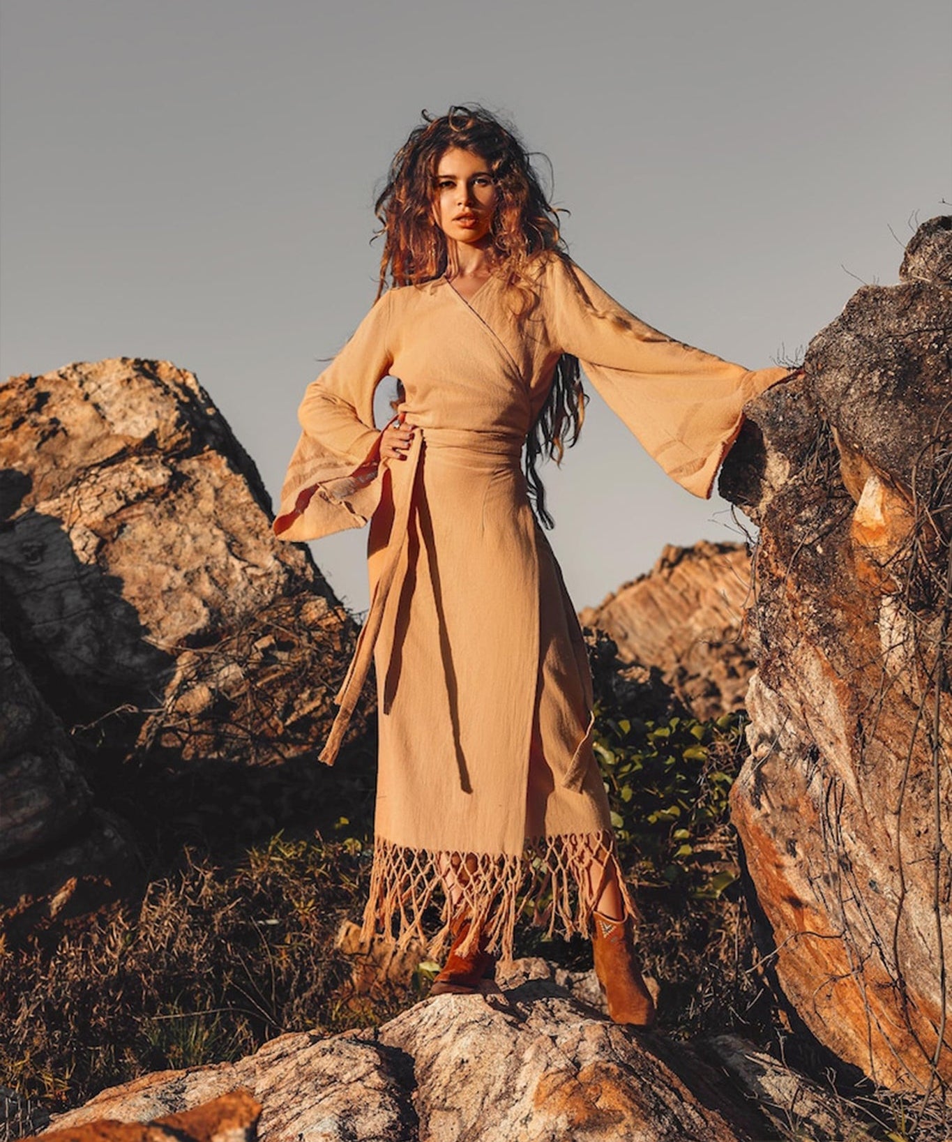 A woman with long, wavy hair stands on rocky terrain, exuding confidence in a tan, long-sleeved dress and boots. She embraces the spirit of ethical fair work production while posing against the rocks under a clear sky, wearing the Boho Maxi Skirt by AYA Sacred Wear — a sustainable cotton skirt that embodies bohemian elegance.