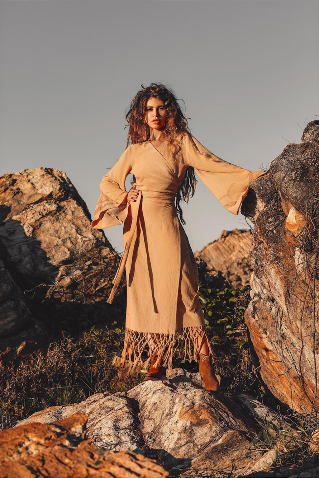 A woman with long, wavy hair stands on rocky terrain, exuding confidence in a tan, long-sleeved dress and boots. She embraces the spirit of ethical fair work production while posing against the rocks under a clear sky, wearing the Boho Maxi Skirt by AYA Sacred Wear — a sustainable cotton skirt that embodies bohemian elegance.