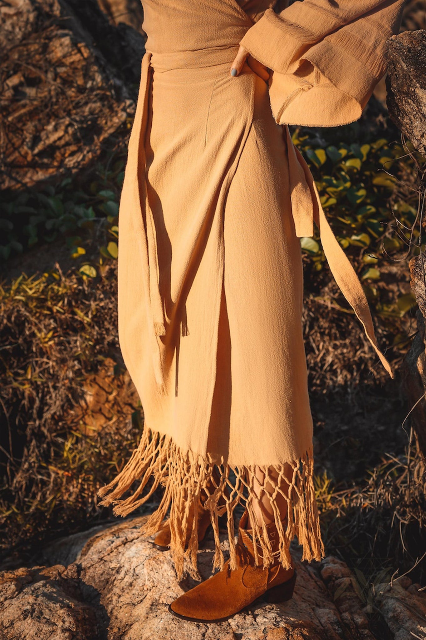 A person is outdoors on a rocky terrain, dressed in an AYA Sacred Wear Boho Maxi Skirt, featuring intricate fringe detailing around the hem, and paired with brown shoes. The lush green foliage provides a backdrop, focusing on the lower half of their body.