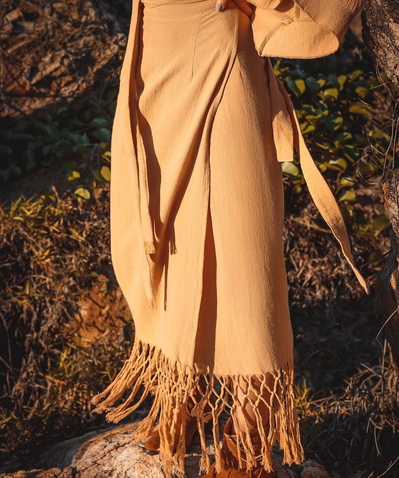 A person is outdoors on a rocky terrain, dressed in an AYA Sacred Wear Boho Maxi Skirt, featuring intricate fringe detailing around the hem, and paired with brown shoes. The lush green foliage provides a backdrop, focusing on the lower half of their body.