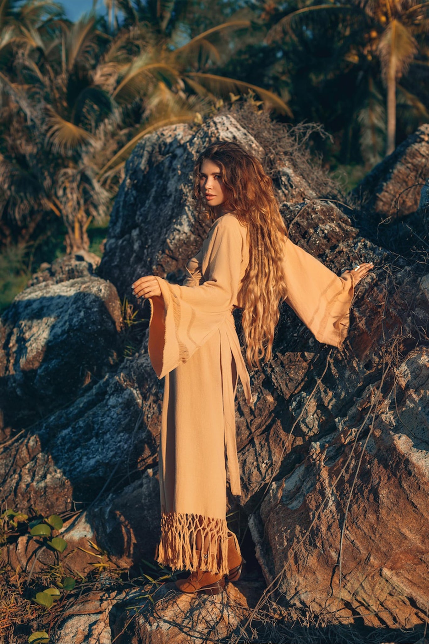 A woman with long, wavy hair wearing a fringed beige dress stands against large rocks in a tropical setting. Surrounded by palm trees and greenery, she exudes effortless style in her AYA Sacred Wear Boho Maxi Skirt, made of sustainable cotton under warm, natural lighting.