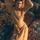 A woman elegantly poses in AYA Sacred Wear's Goddess Skirt—an exquisite Boho Maxi Skirt made from flowing cotton—with eyes gently closed and arms raised against a backdrop of rugged terrain, as the sunlight bathes the scene in warm hues.