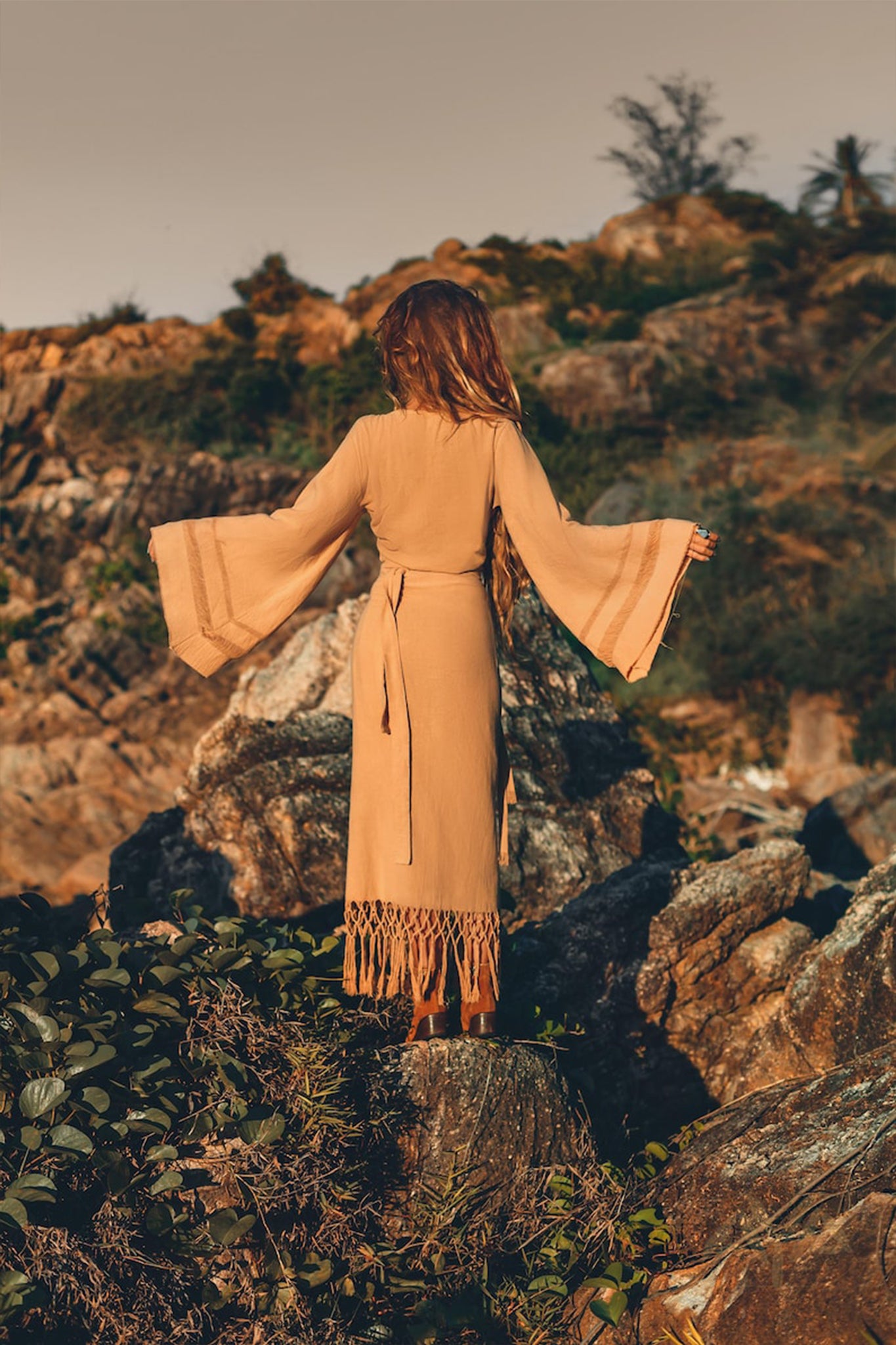 A woman dressed in the AYA Sacred Wear beige Boho Maxi Skirt, adorned with wide sleeves and fringe details, stands gracefully on rocky terrain, exuding a bohemian spirit. Made from sustainable cotton, the skirt perfectly complements the rugged backdrop and sparse greenery beneath a clear sky.