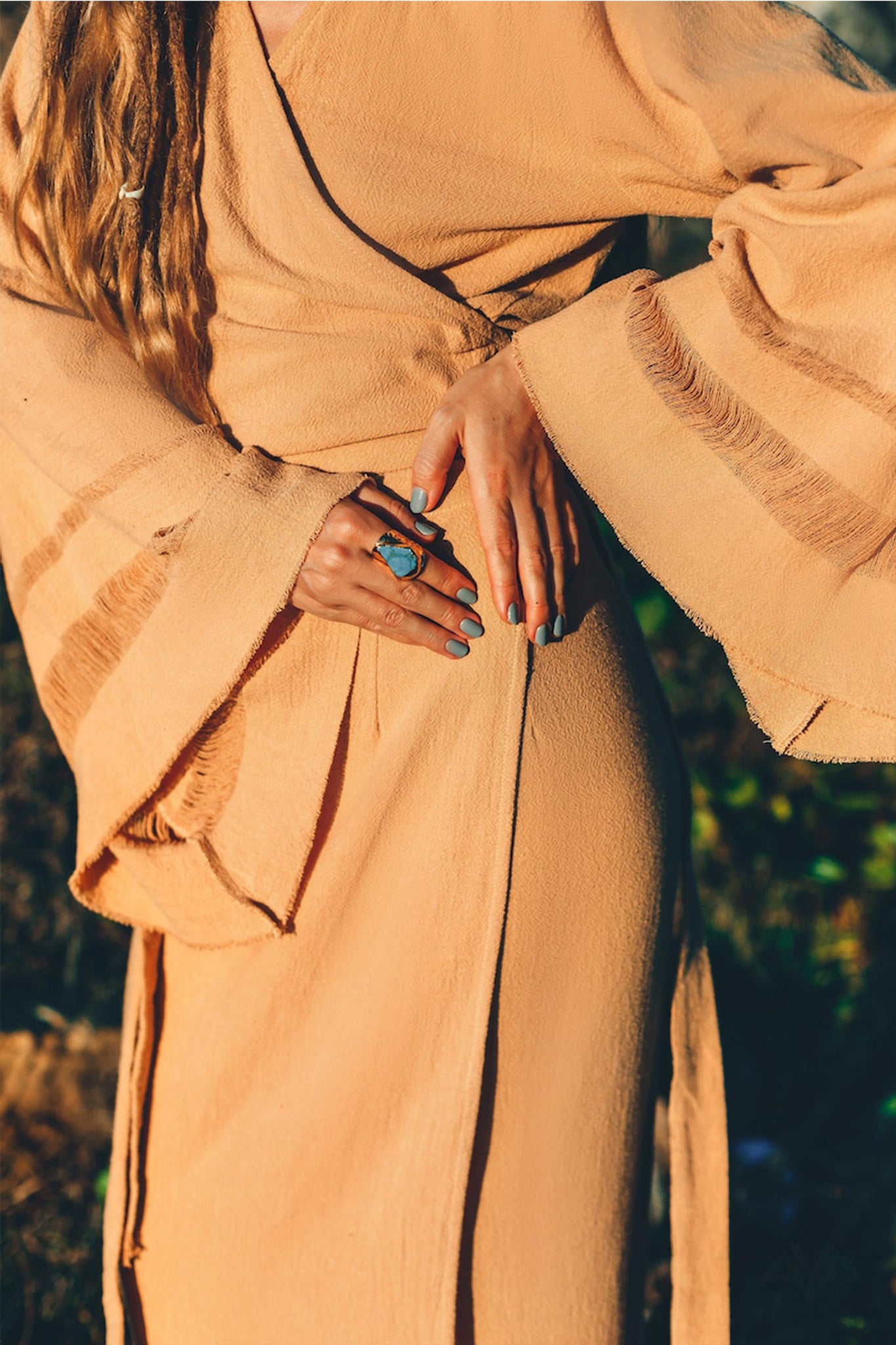 A person wearing a sustainable, textured brown robe with wide sleeves, possibly from the shoulders to the thighs, adorns teal nail polish and a large turquoise ring on their finger. They stand outside in natural sunlight, embodying the bohemian style of AYA Sacred Wear's Boho Maxi Skirt or Goddess Skirt.