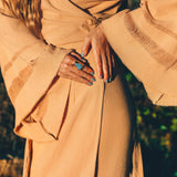 A person wearing a sustainable, textured brown robe with wide sleeves, possibly from the shoulders to the thighs, adorns teal nail polish and a large turquoise ring on their finger. They stand outside in natural sunlight, embodying the bohemian style of AYA Sacred Wear's Boho Maxi Skirt or Goddess Skirt.