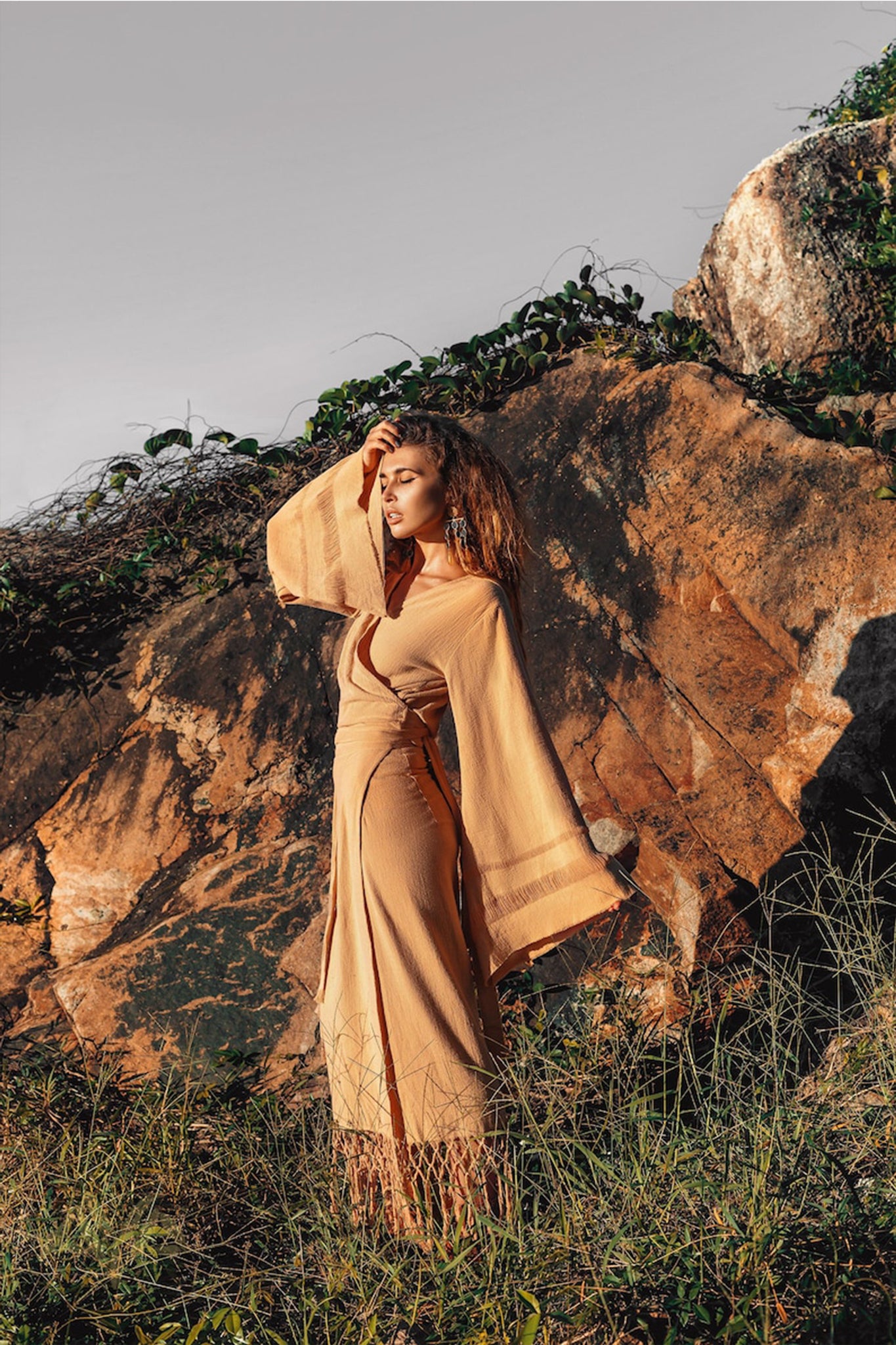 A woman dressed in a long, flowing beige Goddess Skirt by AYA Sacred Wear stands in front of a large rock, gently touching her hair. Surrounded by grass and vines, the outdoor scene is softly illuminated by the overcast sky.