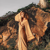 A woman dressed in a long, flowing beige Goddess Skirt by AYA Sacred Wear stands in front of a large rock, gently touching her hair. Surrounded by grass and vines, the outdoor scene is softly illuminated by the overcast sky.