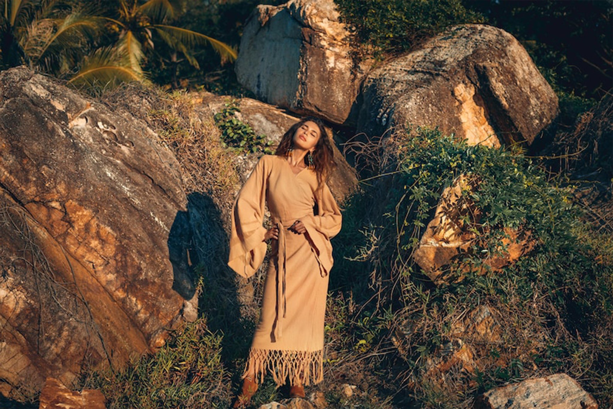 Dressed in a tan outfit and the Boho Maxi Skirt by AYA Sacred Wear, a person stands amidst rugged rocks and greenery, basking in the warm sunlight. With eyes closed and wavy hair, they create a serene, earthy scene that embodies relaxation.