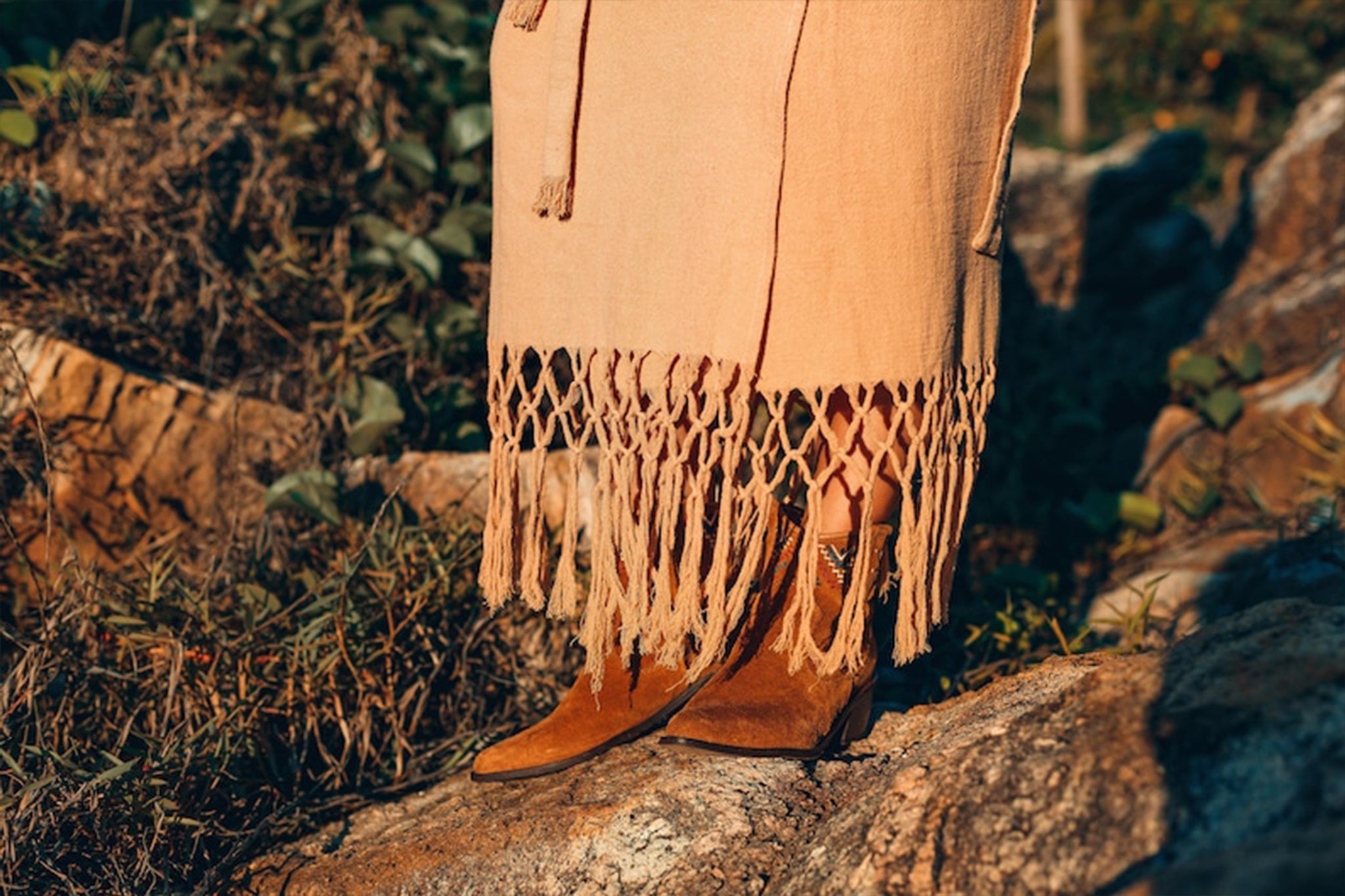 A person wearing brown suede boots and the AYA Sacred Wear Boho Maxi Skirt stands on rocky terrain surrounded by greenery. The image captures the lower half of the outfit in natural sunlight, showcasing its free-spirited elegance.