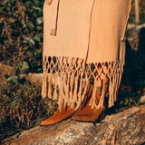 A person wearing brown suede boots and the AYA Sacred Wear Boho Maxi Skirt stands on rocky terrain surrounded by greenery. The image captures the lower half of the outfit in natural sunlight, showcasing its free-spirited elegance.