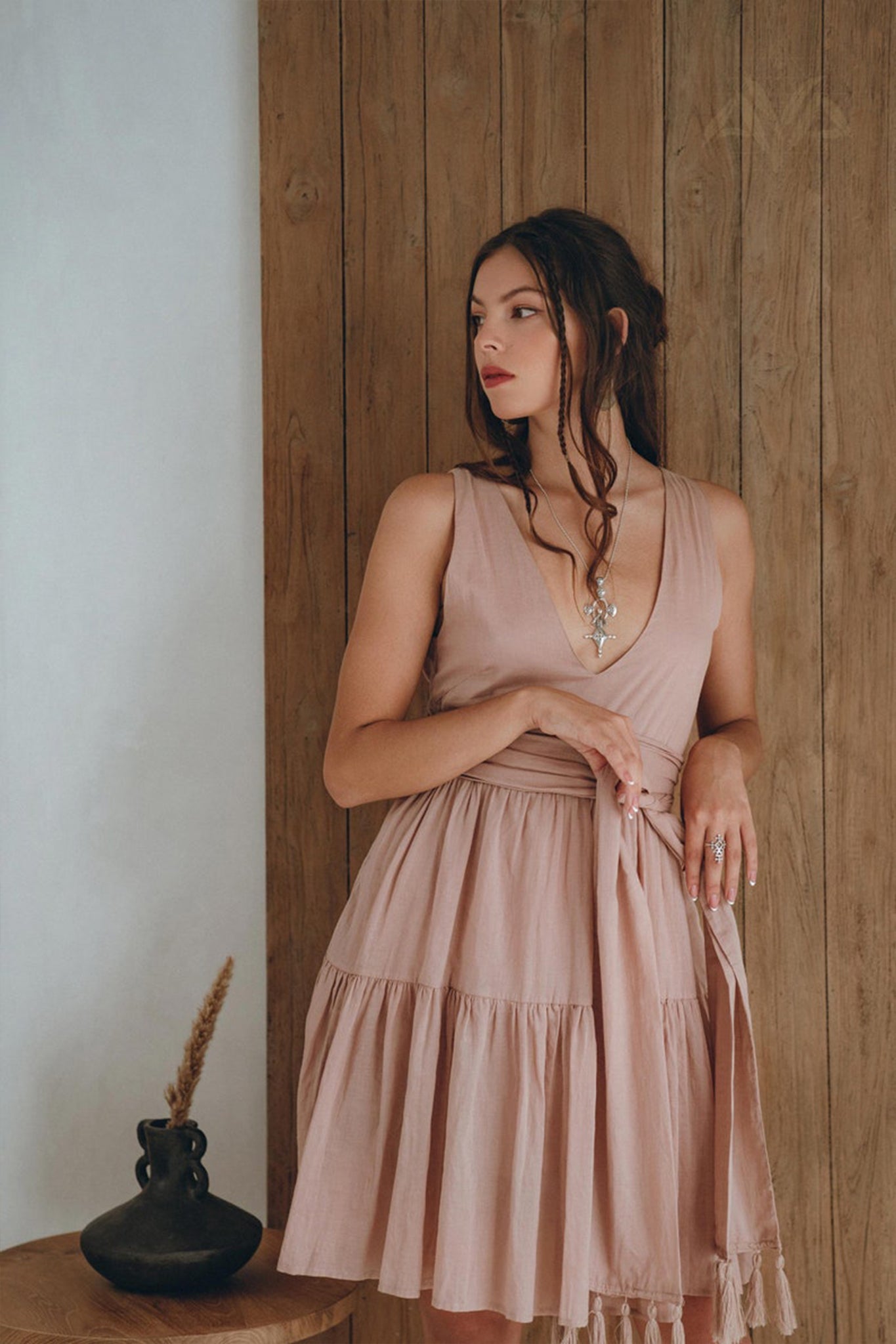 Wearing AYA Sacred Wear's Boho Mini Bridesmaid Dress in dusty pink, a woman stands against a wooden wall. Her hair flows in loose waves as she gazes to the side. Nearby, a black vase filled with dried grass on a small wooden table enhances the boho charm of the scene.