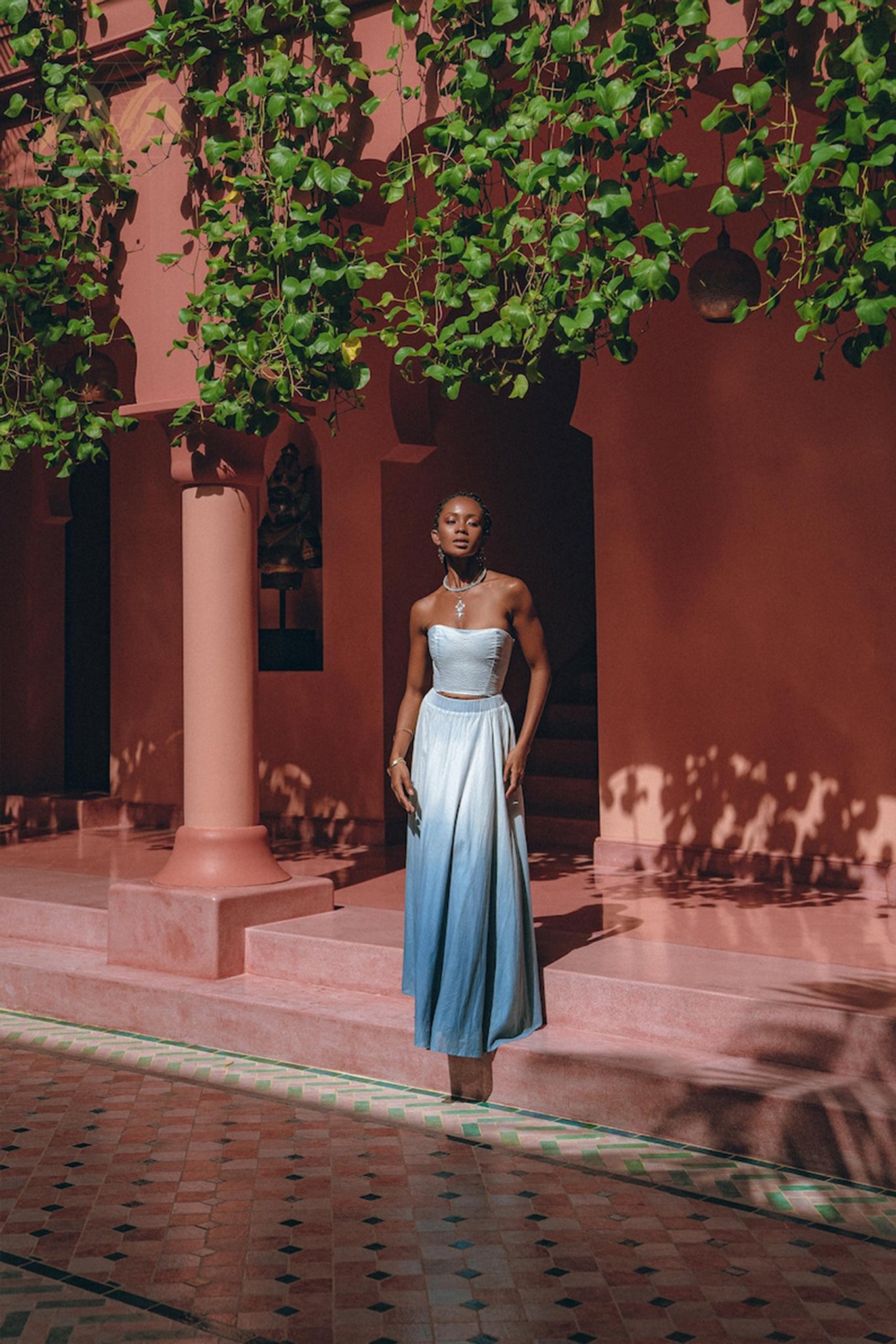 A person stands on a pink tiled patio, enveloped by lush greenery and pink architecture. They are donning the Boho Top Corset Skirt Set from AYA Sacred Wear, an elegant hand-painted blue and white bohemian crop top corset paired with a skirt and matching jewelry, basking in the soft sunlight that filters through the leaves.