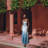 A person stands on a pink tiled patio, enveloped by lush greenery and pink architecture. They are donning the Boho Top Corset Skirt Set from AYA Sacred Wear, an elegant hand-painted blue and white bohemian crop top corset paired with a skirt and matching jewelry, basking in the soft sunlight that filters through the leaves.