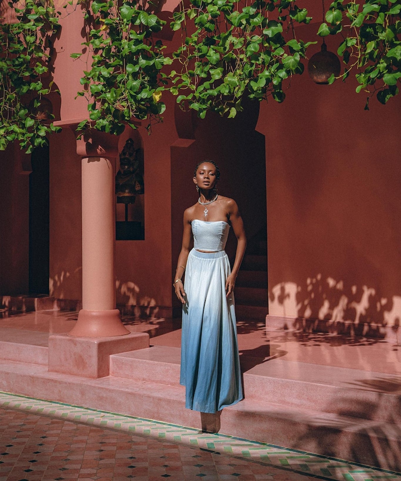 A person stands on a pink tiled patio, enveloped by lush greenery and pink architecture. They are donning the Boho Top Corset Skirt Set from AYA Sacred Wear, an elegant hand-painted blue and white bohemian crop top corset paired with a skirt and matching jewelry, basking in the soft sunlight that filters through the leaves.