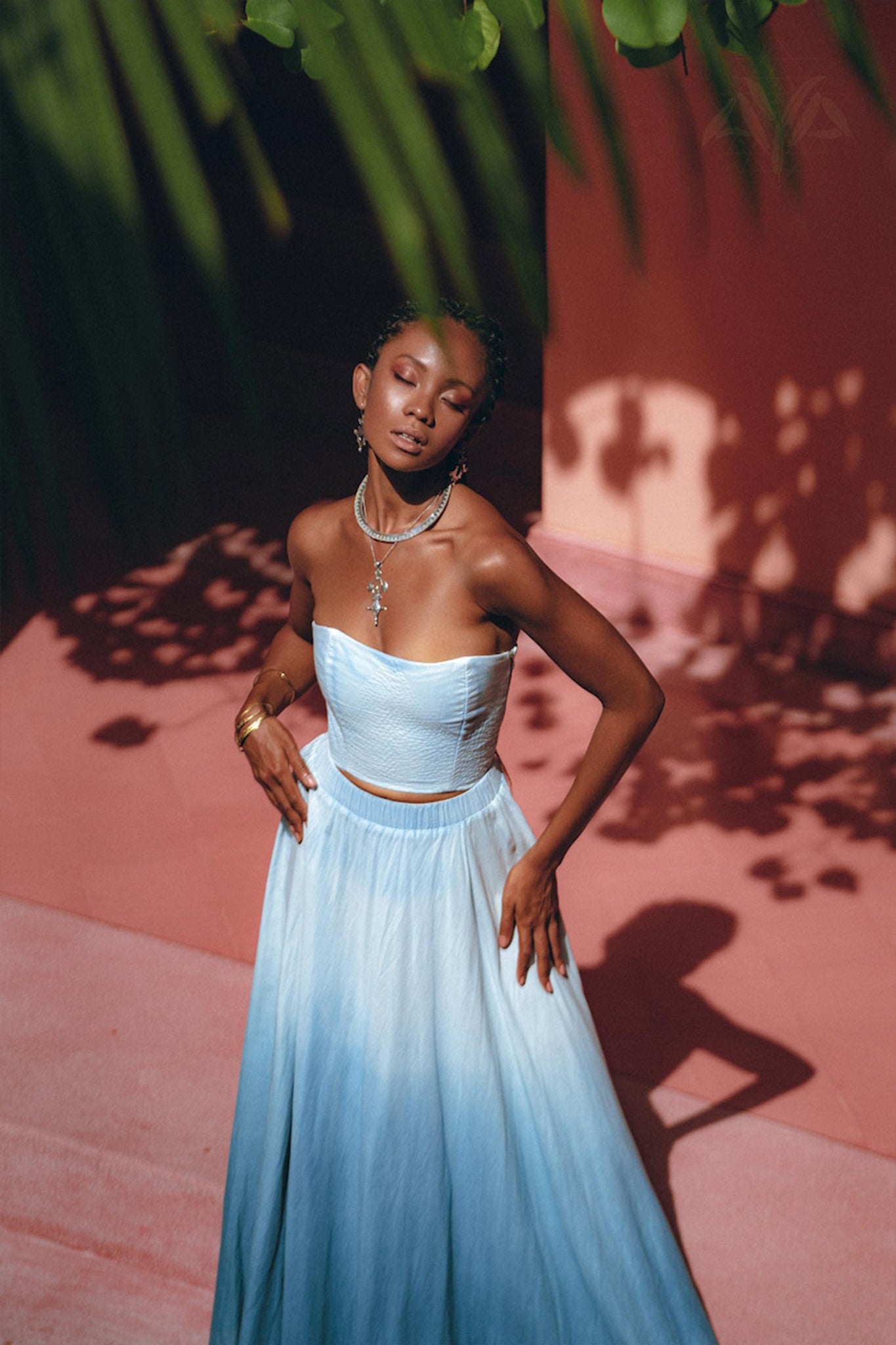 A woman stands gracefully in the Boho Top Corset Skirt Set by AYA Sacred Wear, eyes closed, as sunlight casts leaf shadows around her. The backdrop of a pinkish wall and green foliage above enhances the serene, dreamy atmosphere.