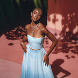 A woman stands gracefully in the Boho Top Corset Skirt Set by AYA Sacred Wear, eyes closed, as sunlight casts leaf shadows around her. The backdrop of a pinkish wall and green foliage above enhances the serene, dreamy atmosphere.